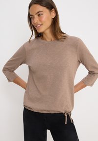 Femme portant un haut taupe à manches longues avec un ourlet à cordon et un pantalon noir, souriante et regardant sur le côté devant un fond blanc uni.