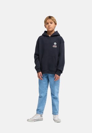 Boy wearing a black hooded sweatshirt with a small logo, light blue jeans, and white high-top sneakers standing against a plain background.