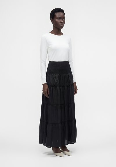 Femme debout portant un haut blanc à manches longues, une jupe longue noire à volants et des chaussures blanches pointues sur un fond blanc uni.