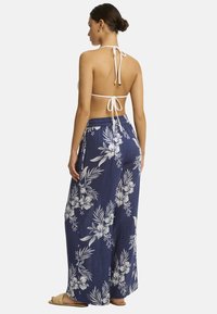 Een vrouw staat met haar rug naar de kijker, gekleed in een witte haltertop en een marineblauwe broek met bloemenpatroon, met een casual kapsel.