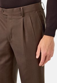 Brūnas, pielāgotas bikses, kas izgatavotas no gludas auduma, ar ģeometrisku priekšpusi, slīpām kabatām un pogu aizdari jostasvietā.