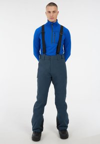 Blauwe langemouwen top met ritskraag, gecombineerd met donkergrijze snowpants met bretels, ritszakken en zwarte accenten bij de enkels.