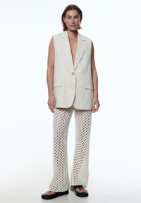 Beige mouwloze blazer met twee voorzakken en een enkele knoop, gecombineerd met losse, witte, gebreide haakbroek en zwarte platform sandalen.