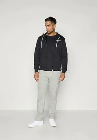 Schwarze Zip-Hoodie mit weißen Kordeln, getragen über einem schwarzen Shirt, kombiniert mit hellgrauen Jogginghosen und weißen Sneakers.