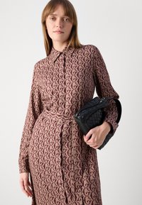 Abito camicia marrone a fantasia con colletto, chiusura con bottoni e tessuto texture. Borsa nera intrecciata tenuta in mano.