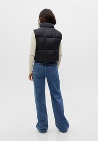 Schwarze, cropped Pufferweste mit hohem Kragen und gesteppten Elementen. Kombiniert mit blauen Straight-Leg-Jeans. Glatte Textur.