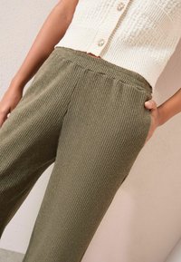Olīvu zaļas, ribotās platās bikses ar mīkstu tekstūru, ar elastīgu jostasvietu un sānu kabatām, apvienotas ar krēma krāsas adītu blūzi.