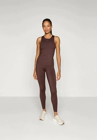 Καφέ αθλητική φανέλα με ταιριαστές κολάν, λεία υφή, εφαρμοστό σχέδιο. Μοντέλο που φοράει λευκά αθλητικά παπούτσια.