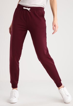 Personne portant un pantalon de jogging bordeaux avec un cordon blanc et des baskets blanches, debout jambes écartées sur un fond uni.