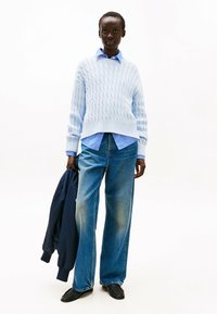 Maglione azzurro a trecce sopra una camicia a righe blu, abbinato a jeans in denim blu a gamba larga e mocassini neri. Tiene in mano una giacca blu navy.