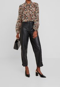 Frau trägt eine gemusterte Bluse mit langen Ärmeln, hochtaillierte schwarze Lederhosen, schwarze Slingback Schuhe mit Blockabsatz und hält eine schwarze Handtasche.