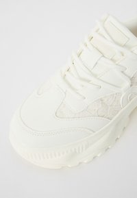 Baskets blanches épaisses avec un panneau de lacets texturé, une surface mate lisse et des lacets plats. Comprend une épaisse semelle en caoutchouc striée.