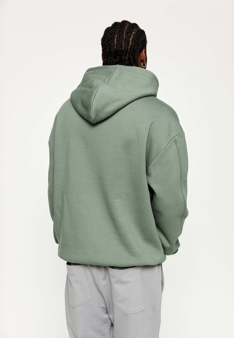 Sudadera verde con capucha, de corte relajado, textura suave y un bolsillo canguro. Pantalones grises con bolsillos visibles debajo de la sudadera.