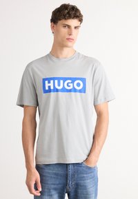 Pilkas medvilninis marškinėliai su mėlynu stačiakampiu logotipu, kuriame užrašyta "HUGO" baltais rašmenimis. Trumpomis rankovėmis ir apvaliu kaklu.