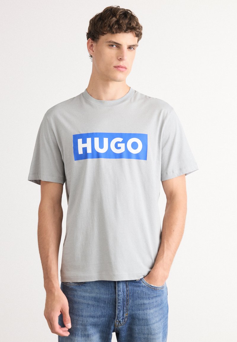 Harmaa puuvillainen t-paita, jossa on sininen suorakulmainen logo ja valkoiset kirjaimet "HUGO". Lyhyet hihat ja pyöreä pääntie.