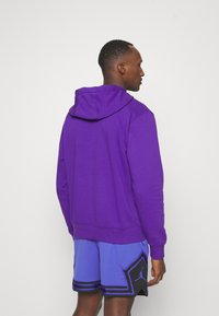 Felpa viola con cappuccio e lacci, texture morbida e polsini a coste. Indossata con pantaloni corti sportivi in rete blu con dettagli neri.