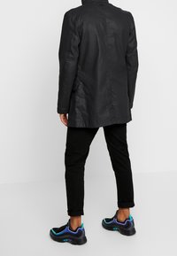Cappotto di pelle nera con design diritto, abbinato a pantaloni neri arrotolati e sneaker neri con accenti blu e viola.
