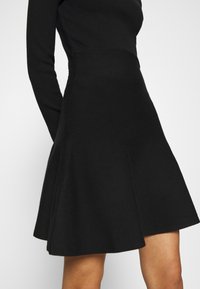 Abito a maglia nera con corpetto aderente, maniche lunghe e gonna svasata. Texture liscia con una leggera lucentezza, le cuciture sono rifinite con cura.