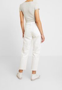 Žena oblečená v přiléhavém šedém tričku s krátkým rukávem, bílé zkrácené džíny s vysokým pasem a rozstřihanými okraji, a bílé vysoké tenisky, stojící zády.