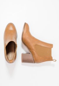 Botines tobilleros marrones con un upper de cuero suave, paneles laterales elásticos y un tacón de bloque texturizado. Incluyen una pequeña pestaña para facilitar su calzado.