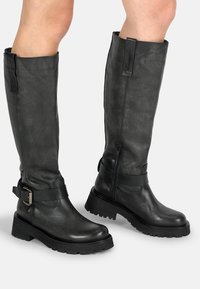 Bottes en cuir noir montantes avec une finition texturée, fermeture éclair sur le côté et détails de sangle à boucle. Semelle épaisse à traction.