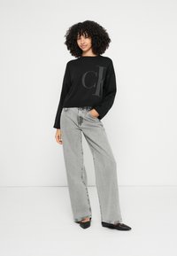 Melns džemperis ar CK logotipu, brīva piegriezuma, ar ribotu apakšu. Kombinēts ar augsta vidukļa gaiši pelēnām platu kāju džinsiem un melnām punktiņveida kurpēm.