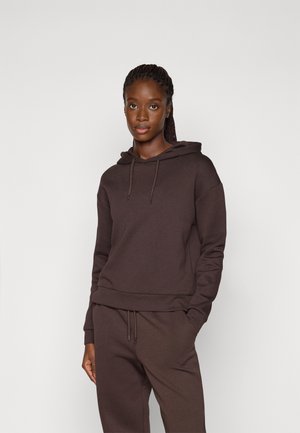 Jeune femme aux cheveux tressés portant un sweat à capuche marron foncé et un pantalon assorti, debout devant un fond clair uni.