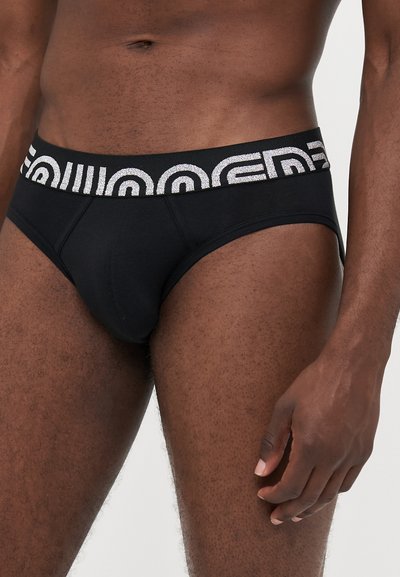 Mandlig torso iført sorte trusser med en sort talje, der har et hvidt geometrisk mønster, stående mod en ensfarvet baggrund.