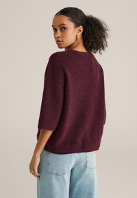 Tumši sarkans oversize adīts džemperis ar pusgarām piedurknēm, reljefu virsmu un plašu kaklu, apvienots ar gaismēji ziliem augsta vidukļa džinsiem.