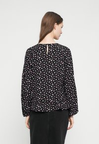 Donna con lunghi capelli castani che indossa una camicetta nera a maniche lunghe con piccole stampe floreali rosa e bianche e una gonna nera, vista da dietro.