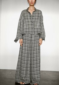 Modelka má na sobě volně střižený šedý kostkovaný outfit, skládající se z košile na knoflíky a širokých kalhot, stojí na tmavé podlaze na světlém pozadí.