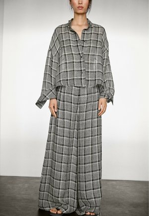 Een model draagt een losvallende, geruite, grijze outfit bestaande uit een overhemd met knopen en wijde broek, staande op een donkere vloer tegen een lichte achtergrond.