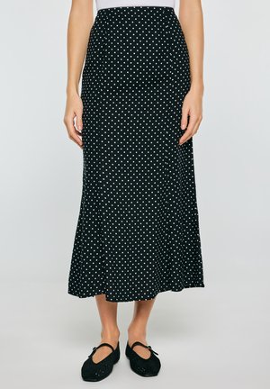 Czarna spódnica midi w białe groszki, noszona z czarnymi butami Mary Jane, pokazana od talii do stóp na jednolitym tle.