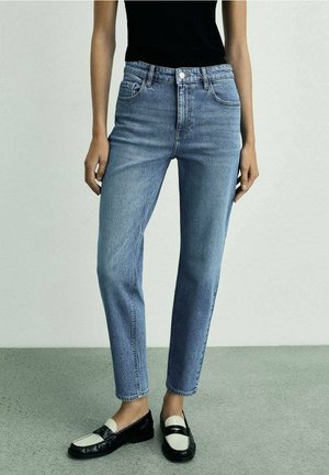 Person trägt hellblaue Jeans mit hoher Taille, schwarz-weiße Loafers und ein schwarzes ärmelloses Oberteil und steht auf einem grauen Boden vor einer schlichten Wand.