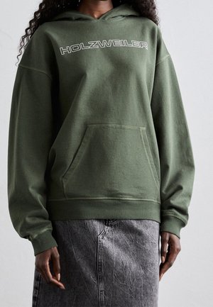 Personne portant un sweat à capuche vert olive avec le texte "HOLZWEILER" sur la poitrine et une poche frontale, associé à une jupe en jean grise.