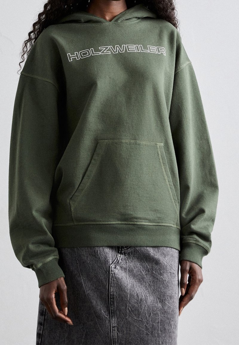 Personne portant un sweat à capuche vert olive avec le texte "HOLZWEILER" sur la poitrine et une poche frontale, associé à une jupe en jean grise.