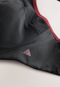 Gros plan sur un vêtement de sport en tissu noir avec une bordure bordeaux, montrant un détail en maille texturée et un petit logo géométrique bordeaux.