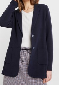 Námořnický blazer vyrobený z texturované tkaniny, s dvěma předními kapsami, dvěma knoflíky a upraveným střihem. Nesený přes bílou žebrovanou košili.