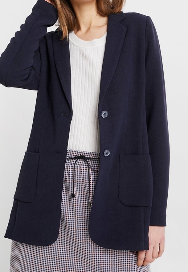 Námořnický blazer vyrobený z texturované tkaniny, s dvěma předními kapsami, dvěma knoflíky a upraveným střihem. Nesený přes bílou žebrovanou košili.