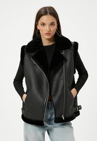 Schwarze Lederweste mit strukturierter Oberfläche, Kunstfellkragen, Reißverschluss und seitlichen Taschen. Akzente sind verstellbare Schnallen an der Taille.