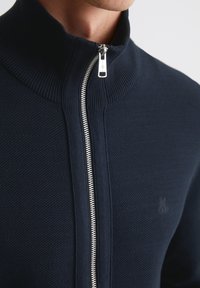 Marineblauwe ritssluitingtrui met een hoge kraag, voorzien van een geribbelde textuur en een zilveren rits. Subtiele logo-detailing aan de voorkant.