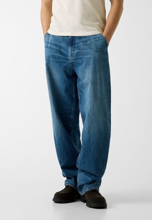 Personne portant un jean bleu ample, les mains dans les poches, un t-shirt blanc et des chaussures décontractées noires, se tenant devant un fond uni.