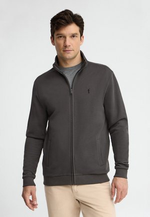 Mężczyzna w ciemnoszarej kurtce na zamek błyskawiczny, na szarej koszuli i beżowych spodniach, stoi na jednolitym jasnym tle.