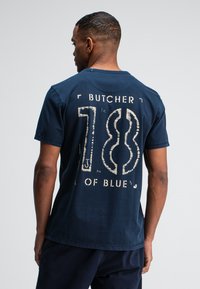 Marineblauwe katoenen T-shirt met een grote witte grafische afbeelding met de tekst "BUTCHER 18 OF BLUE." Ronde hals, korte mouwen, ontspannen pasvorm.