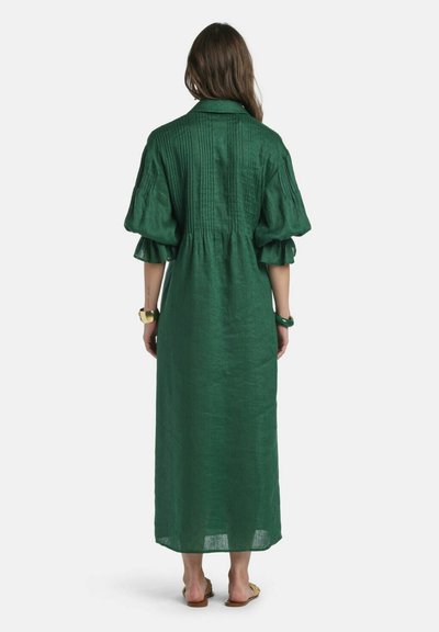 Mujer con cabello largo y castaño que lleva un vestido suelto verde oscuro hasta los tobillos con mangas abullonadas, de pie descalza contra un fondo sencillo.