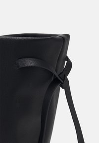 Czarna skórzana torebka o teksturowanym wykończeniu, z detalem w postaci związanej rączki. Kształt jest sztywny z zwężającym się otwarciem u góry.