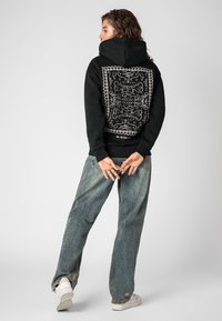 Zwarte hoodie met een ingewikkeld wit paisleymotief op de achterkant. Gecombineerd met loszittende lichtblauwe jeans en witte sneakers. Minimalistische uitstraling.