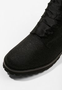 Botín negro de tobillo hecho de material texturizado con un acabado brillante, que presenta volantes a lo largo del lateral y una suela gruesa y robusta.