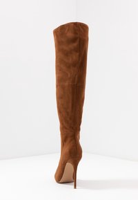 Botte en daim marron au-dessus du genou avec un design épuré, un bout pointu et un talon aiguille haut. Texture lisse et coutures visibles minimales.