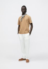 T-shirt de algodão castanho, calças de linho brancas, espadrilles castanhas e um lenço com padrão preto e branco criam um conjunto casual.